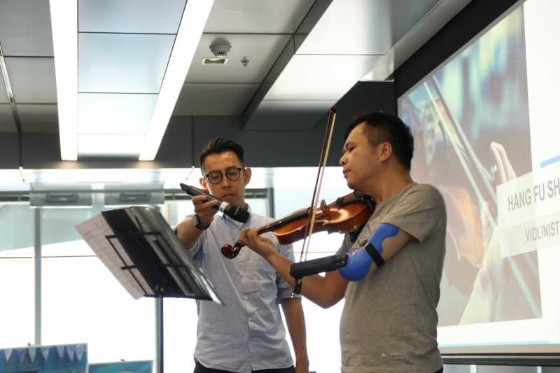 man with prosthetic arm plays violin while another man holds a microphone for his instrument