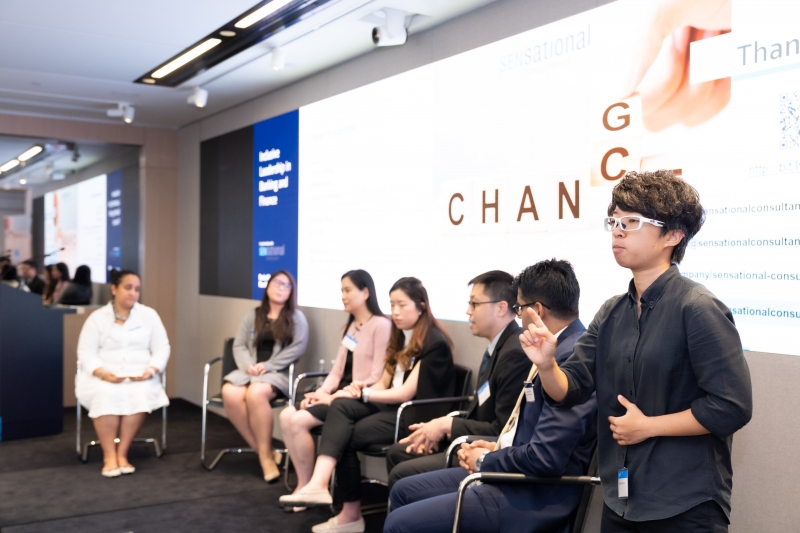 a woman in a blue shirt signs as a panel takes place in the background