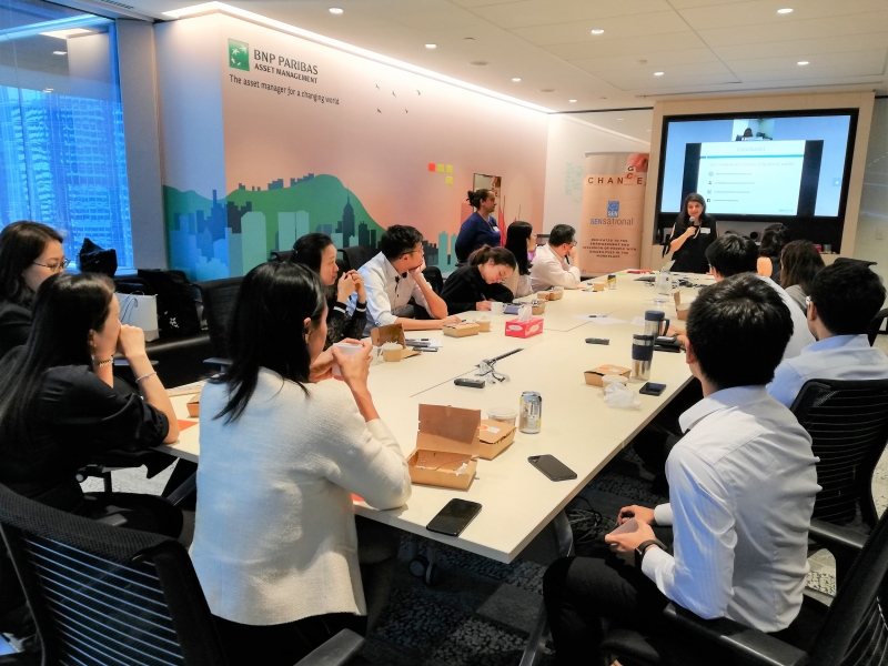 group of women and men in business attire listening attentively to a presentation by a woman standing up