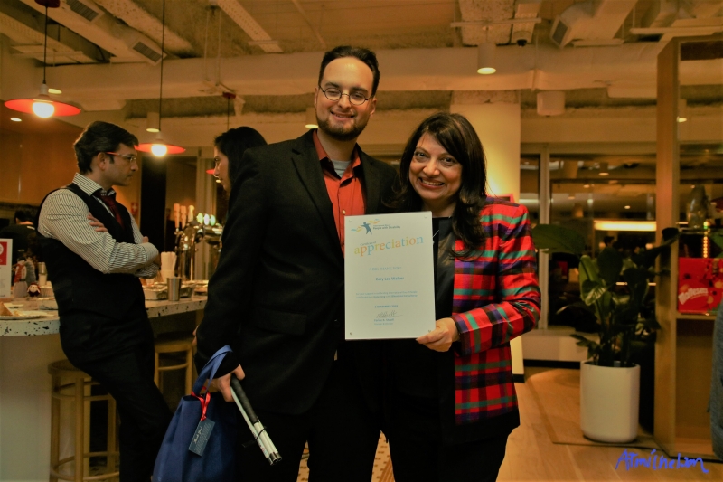 Woman in a colorful boxy jacket holding a certificate stands proudly next to a man with glasses
