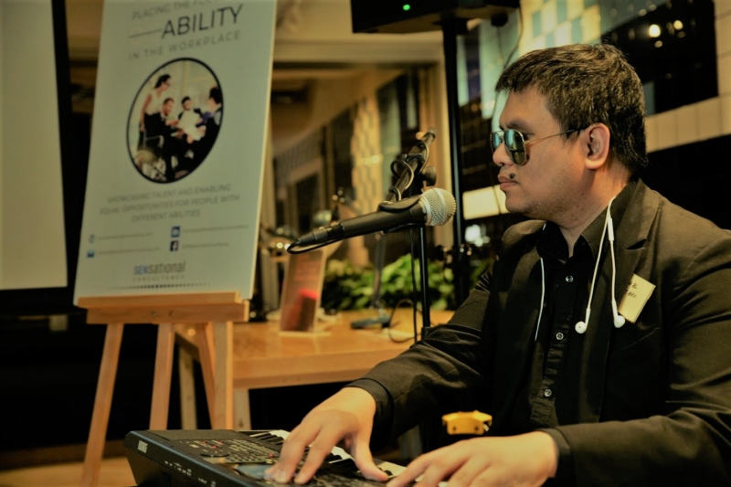 A man wearing dark sunglasses and a black suit jacket, with ear phones draped around his neck, plays the keyboard