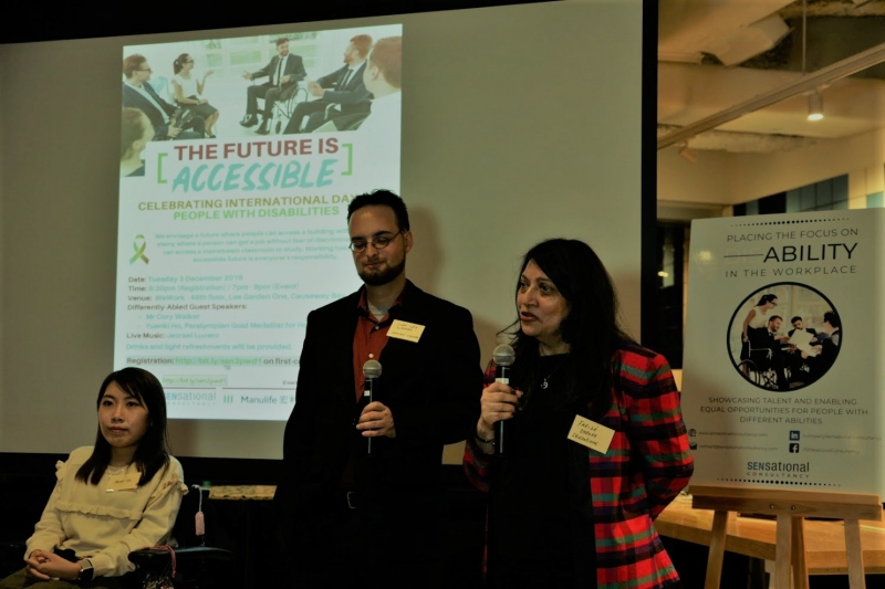 A woman in a colorful boxy jacket speaks into a microphone in front of a screen, with a man and a woman using a wheelchair beside her