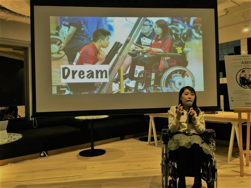 A woman wearing a white dress and using a wheelchair speaks into a microphone in front of a slideshow with the word "dream" and pictures of her athletic career