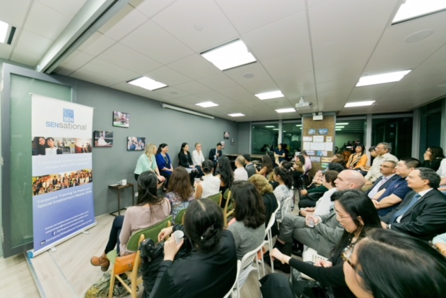 In a group photo of all panelists and audience members, 4 women and a man (panel members) in business attire are sitting at the front facing the audience members who are sitting in rows.