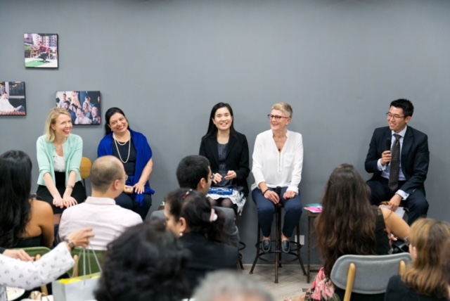4 women and a man (panel members) in business attire are sitting at the front mid-discussion, at the bottom of the picture audience members can be seen sitting in rows.