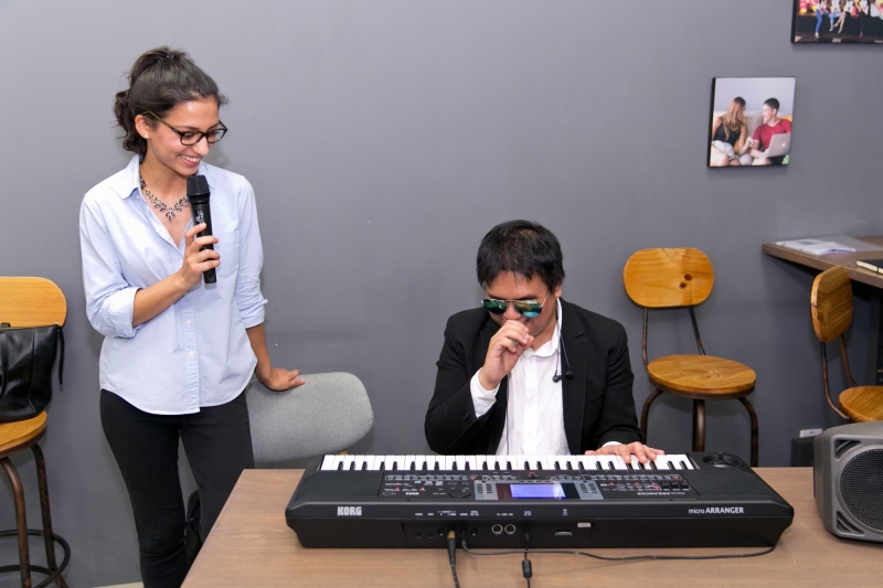 A woman is interviewing the visually impaired musician, the musician is sitting behind his piano whilst the woman is standing next to him.