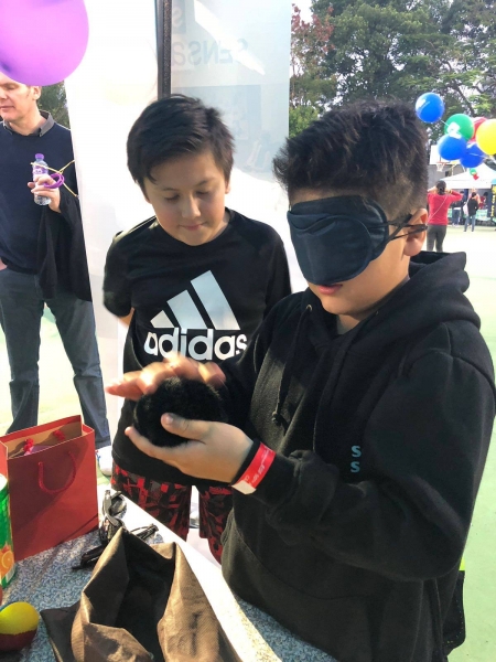 A boy with a blindfold on is feeling a ball and trying to describe what he feels, another boy can be seen watching on the side.