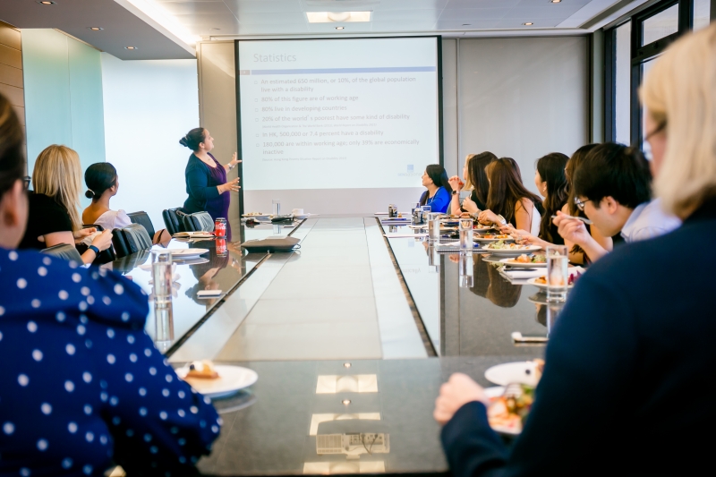 Skadden staff members are seated at a table, paying attention to the presentation given by SENsational staff members whilst eating their lunch.