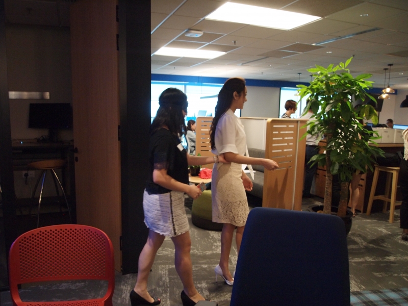 2 Colliers staff participating in a sensory deprivation activity. 1 staff is blindfolded while another staff without a blindfold is helping the blindfolded staff member navigate the surrounding areas.