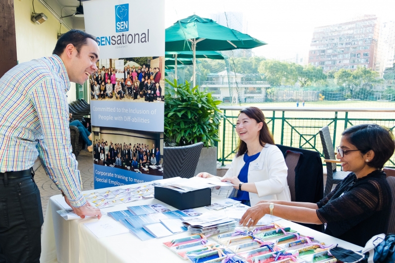 A man in business attire is checking into the event at the SENsational reception desk. To women in business attire, seated behind the desk help him check-in. On the desk are SENsational Foundation brochures