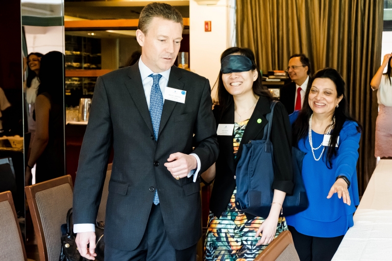 A woman in blindfolds is being guided to take a seat by a man they are followed by the Faride Shroff. All 3 of them are dressed in business attire.
