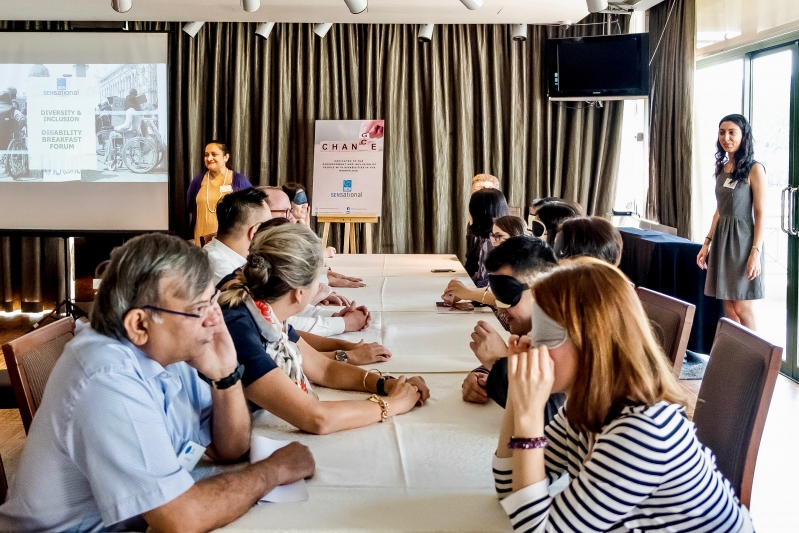 A group of men and women are seated at a table, half of them are blindfolded and the other half are not. Each blindfolded participant is having a conversation with a non-blindfolded participant.