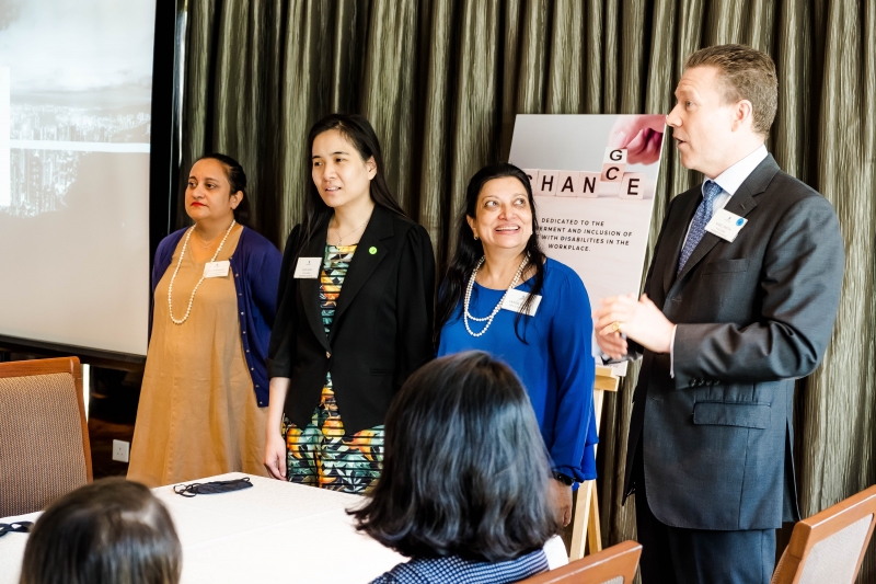 3 women and 1 man are facing the audience members and speaking. They are dressed in business attire