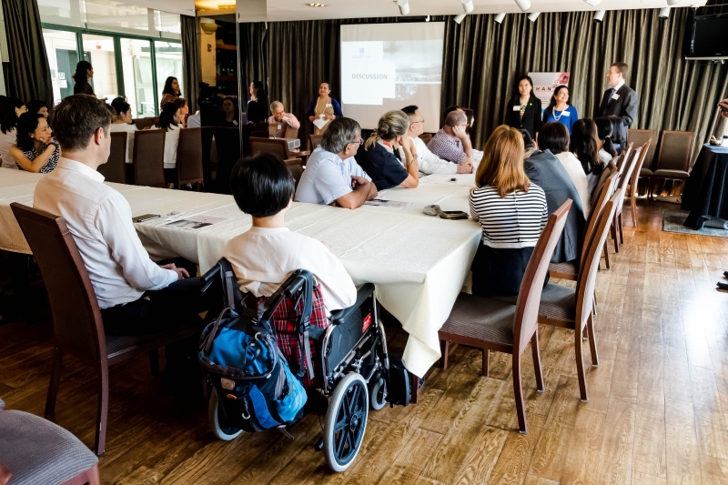 Men and women in business attire are seated by tables whilst listening to 3 women and 1 man speaking. 