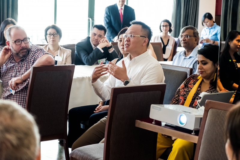 A group of men and women dressed in business attire seated at tables, a man in a white shirt is speaking.